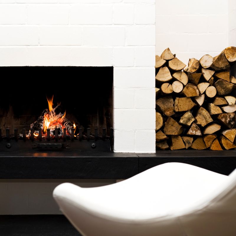 a wood fireplace with white surround by a stack of wood and a white chair